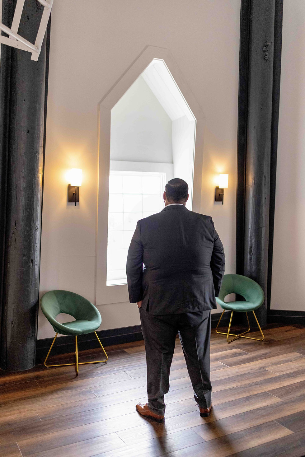 A man in a suit stands facing a large arched window. The room has wooden floors, green chairs, wall lights, and a calm ambiance.
