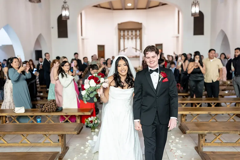 Bride and groom smile, walking down the aisle of a church. She holds red and white bouquet. Guests stand, taking photos. Romantic mood.