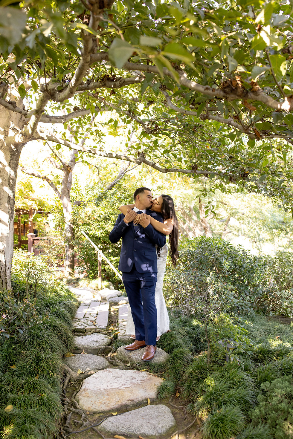 Couple kisses under a leafy canopy on a stone path in a lush garden. The man wears a blue suit; the woman in a white dress embraces him.