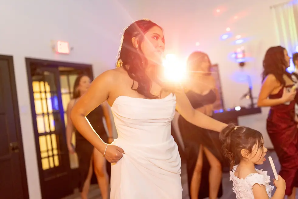Woman in a white dress stands with a child holding a treat. Bright light effect in a festive indoor setting with people in elegant outfits.