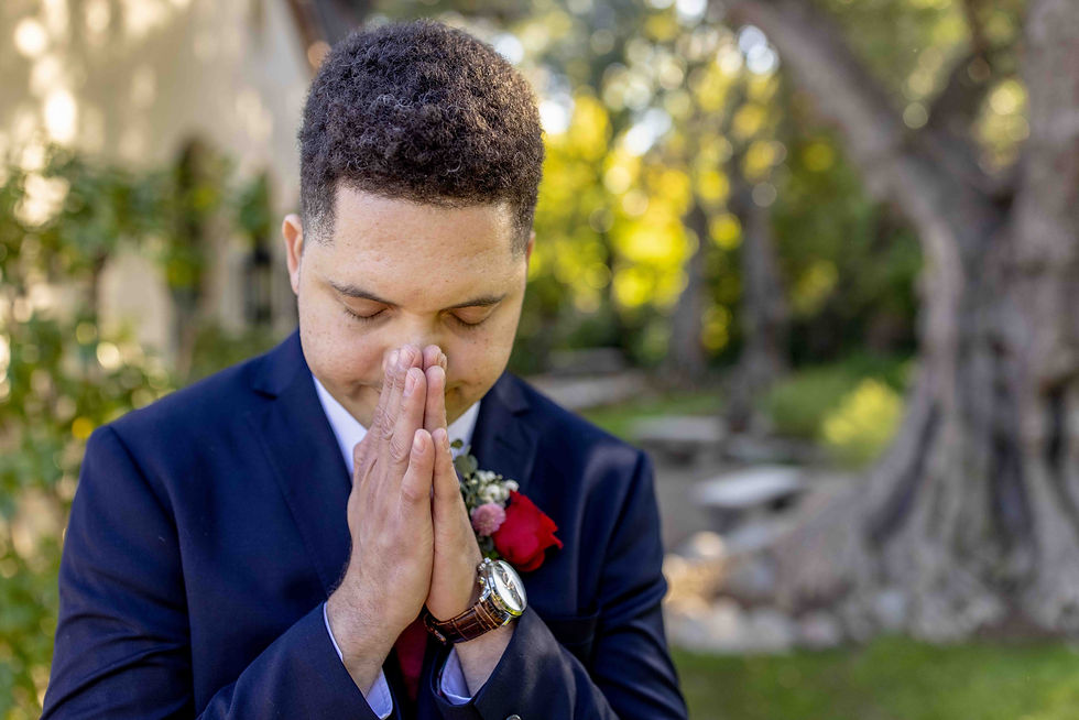 Man in a navy suit with a red rose boutonniere stands outside, eyes closed, hands in a prayer pose. Sunlit trees in the background.