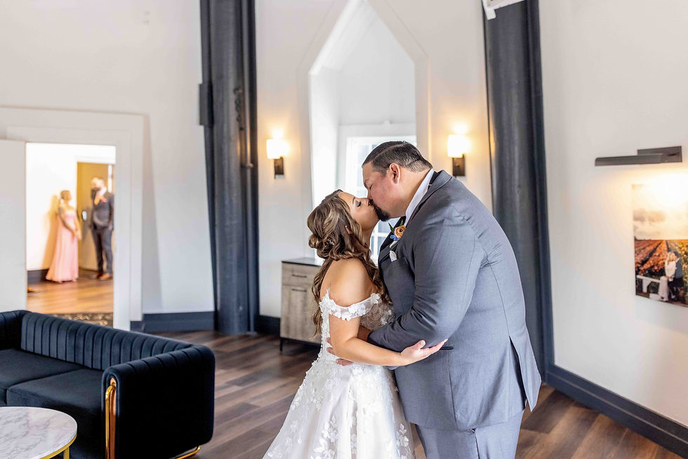 Bride and groom kissing indoors, dressed elegantly. Soft lighting, wood floor, and a modern sofa. Two guests are visible in the background.