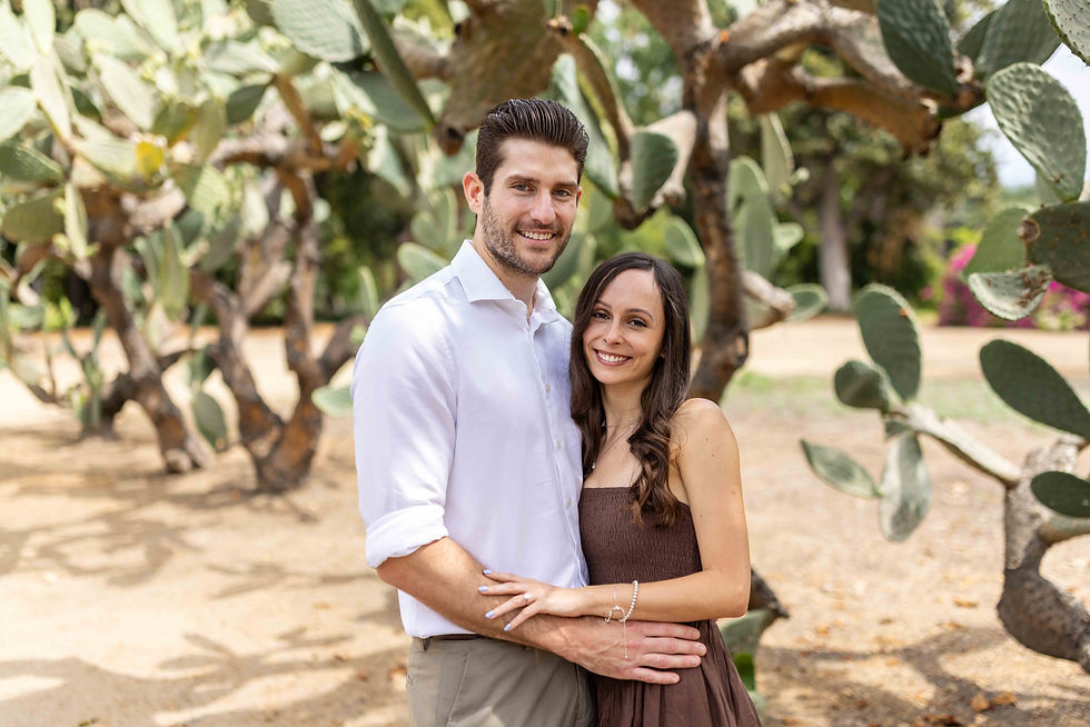 Couple smiling and embracing in a sunny garden with cactus plants around. The woman wears a brown dress; the man, a white shirt.