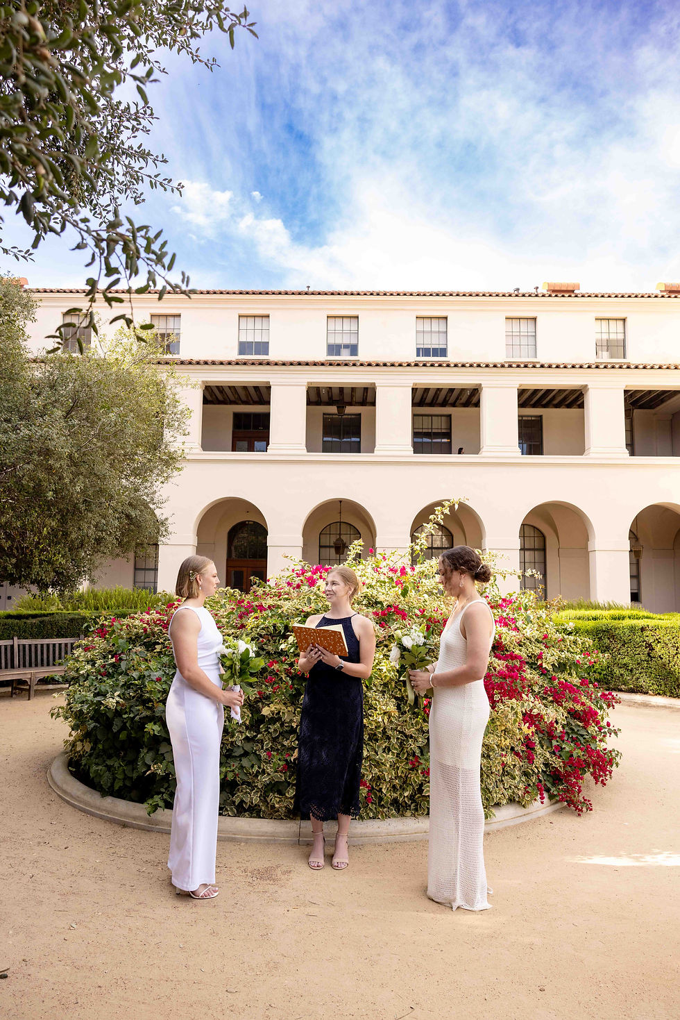 Three women in formal attire stand by flowers, one holding a book. Background shows an elegant building and blue sky, suggesting a ceremony. Pasadena City Hall Same Sex wedding