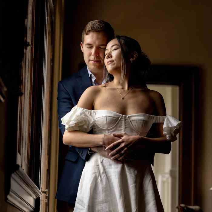 A couple embraces near a window. The woman in a white dress closes her eyes, while the man in a blue suit gently hugs her. Warm lighting.