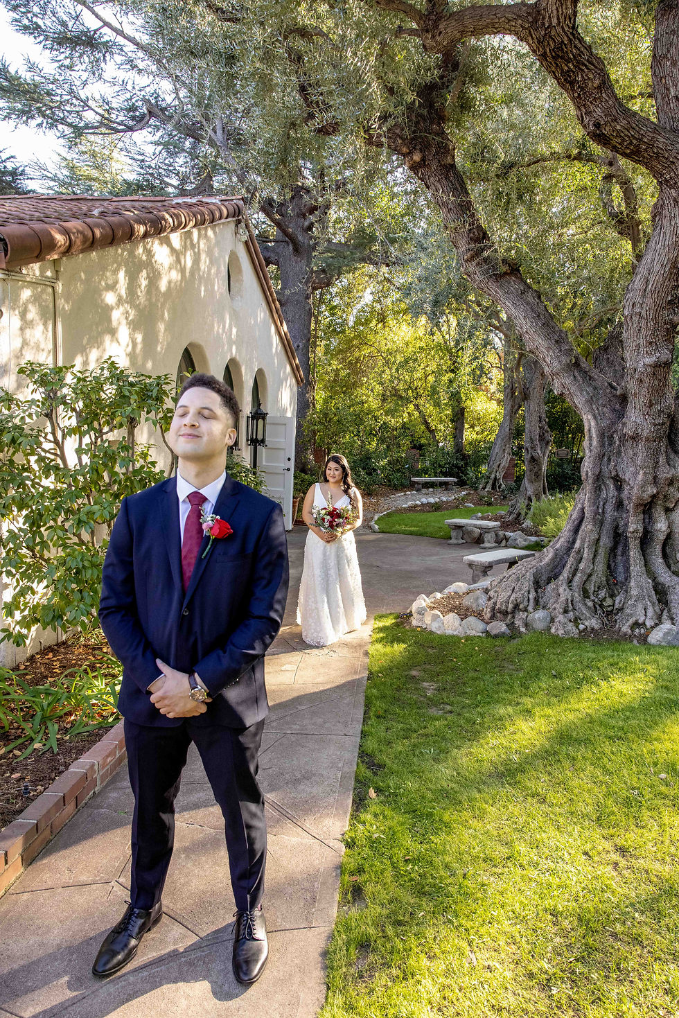 Groom in navy suit with red rose stands smiling with eyes closed. Bride in white dress approaches from behind. Outdoor garden scene.