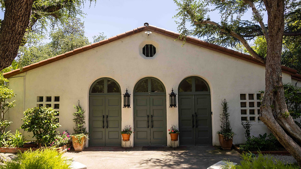 Stucco building with three green doors, arched windows, and lanterns. Surrounded by lush trees and potted plants, creating a serene atmosphere.