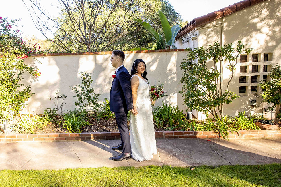 Bride and groom standing back to back, holding hands in a garden. Bride holds bouquet. Sunlit wall and foliage in the background.