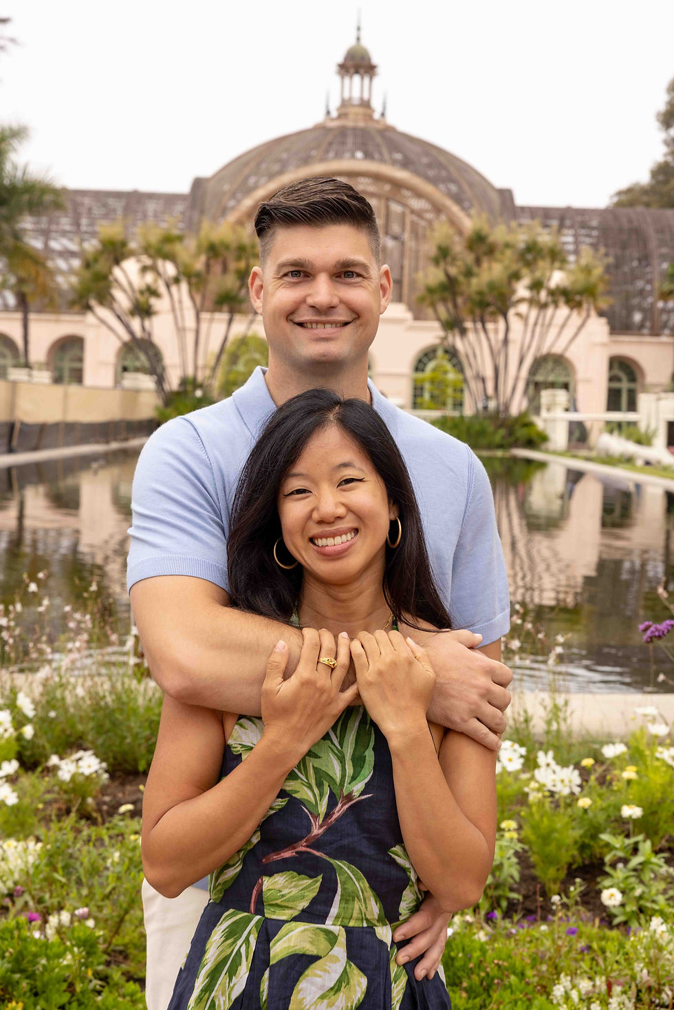 Smiling couple embracing in a garden with a historic domed building in the background. Bright floral dress, light blue shirt, peaceful setting.