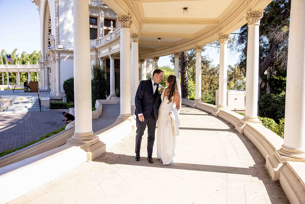 Bride and groom share a kiss under elegant white columns of a grand building. Sunlight casts shadows, greenery in the background. Romantic mood.