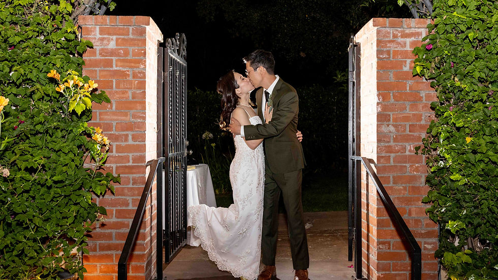 A bride and groom kiss under an archway at night. She wears a white gown; he wears a dark suit. Brick pillars and lush greenery frame them.