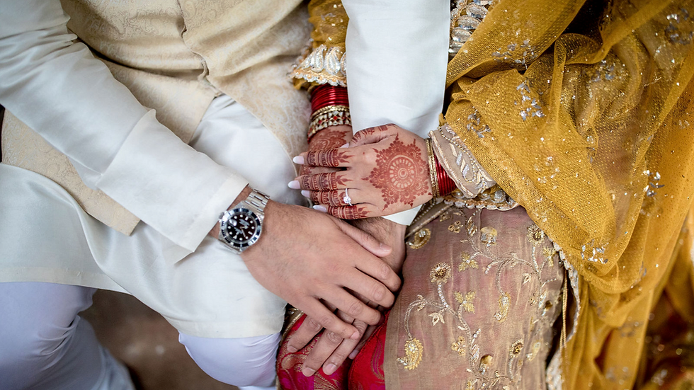 Close-up of a couple's hands resting on each other in ornate attire. Henna on the woman’s hand, gold and white attire, a watch visible.