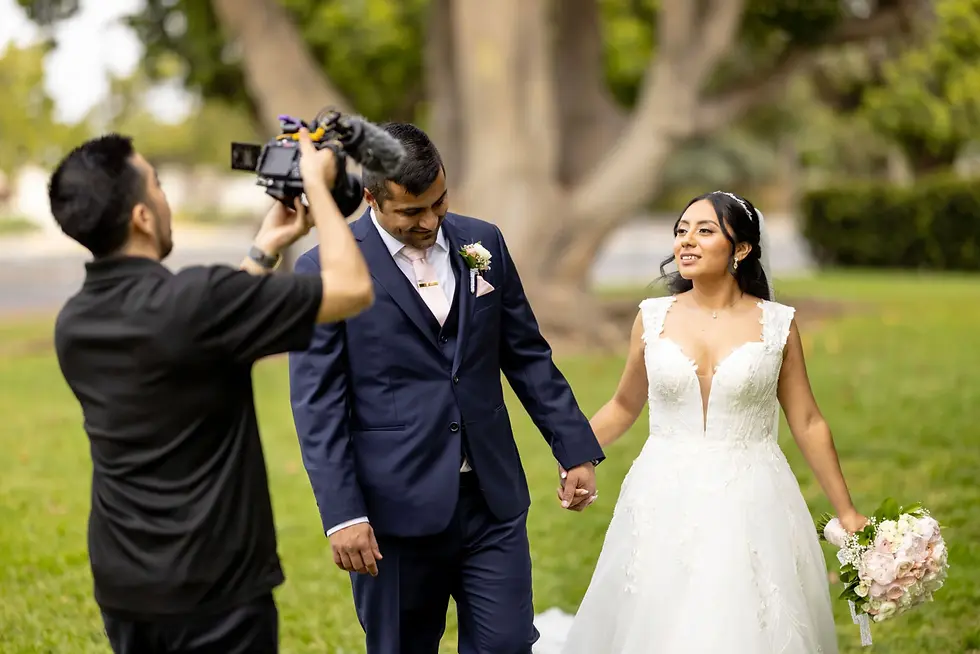 Bride and groom hold hands, smiling, as cameraman films them in a park. Bride wears a white gown, groom in a navy suit. Lush greenery.