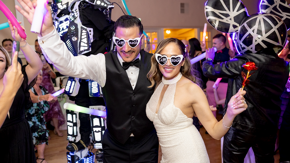 Couple dancing at a party, wearing heart-shaped glasses with "E & M 08.24.24". Neon lights and costumed performers in the background.