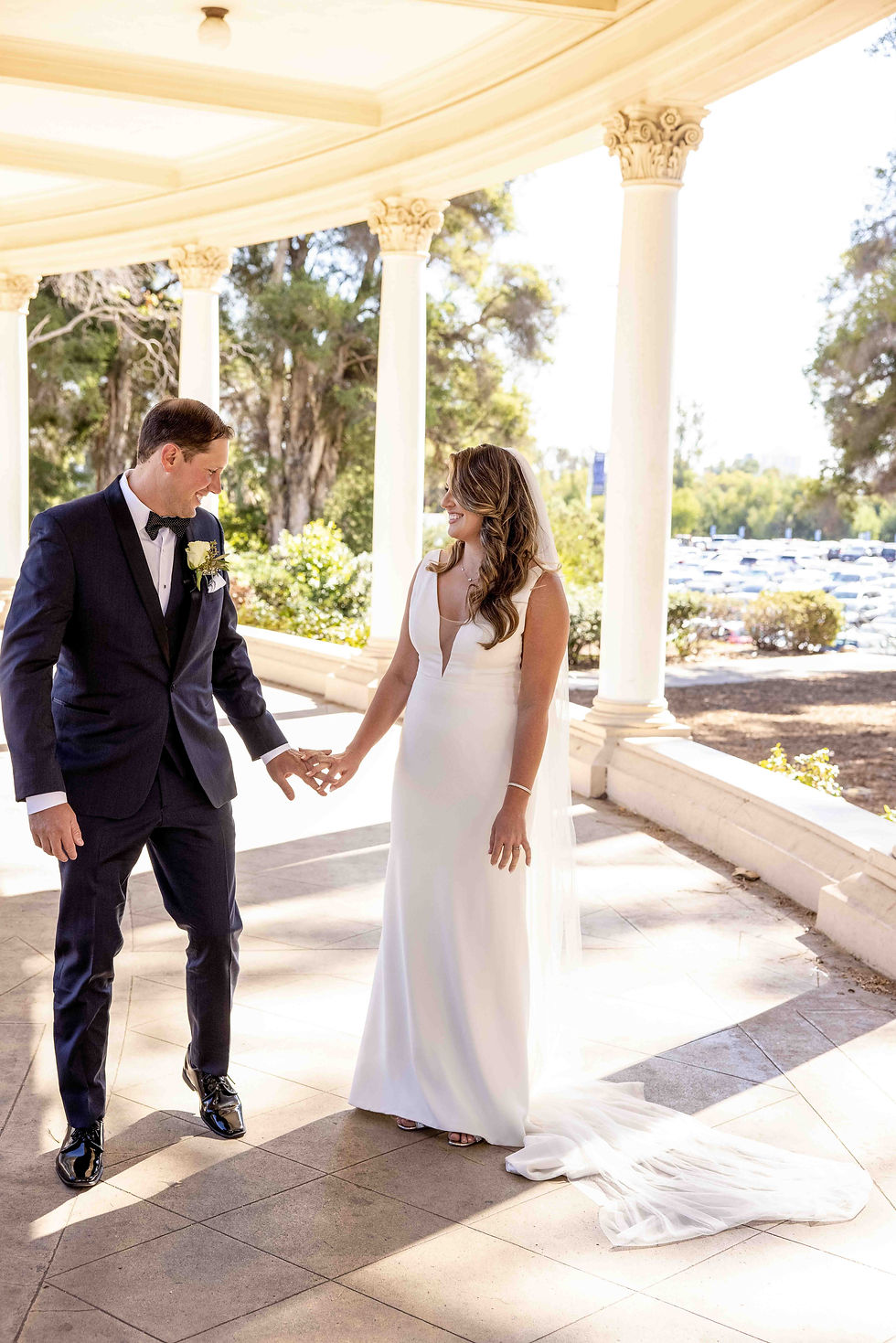 Bride in white gown and groom in suit hold hands, smiling at each other under a sunlit pavilion with trees in the background.