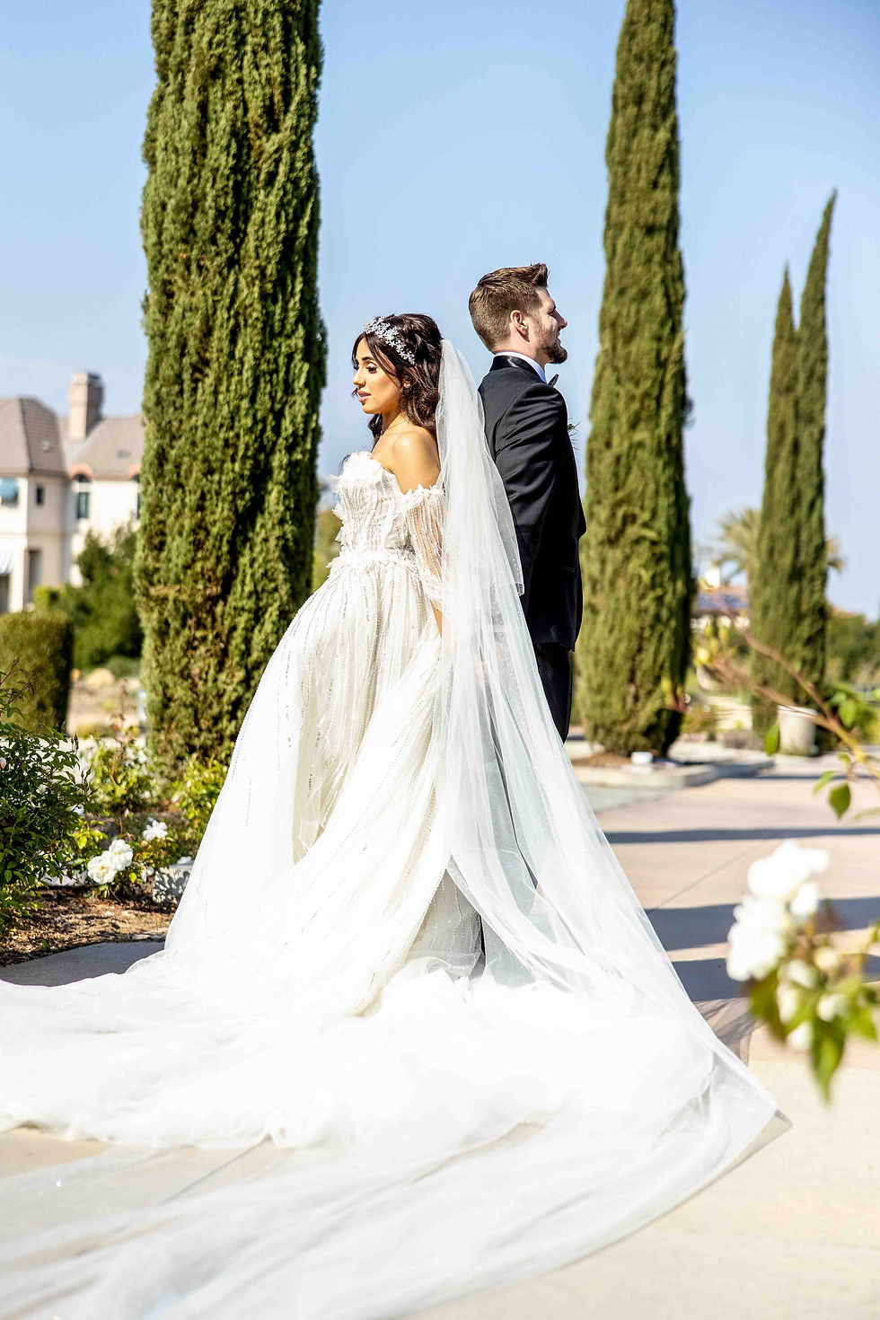Bride in white gown and groom in black tux stand back-to-back outdoors, surrounded by tall trees and flowers, under a clear blue sky.