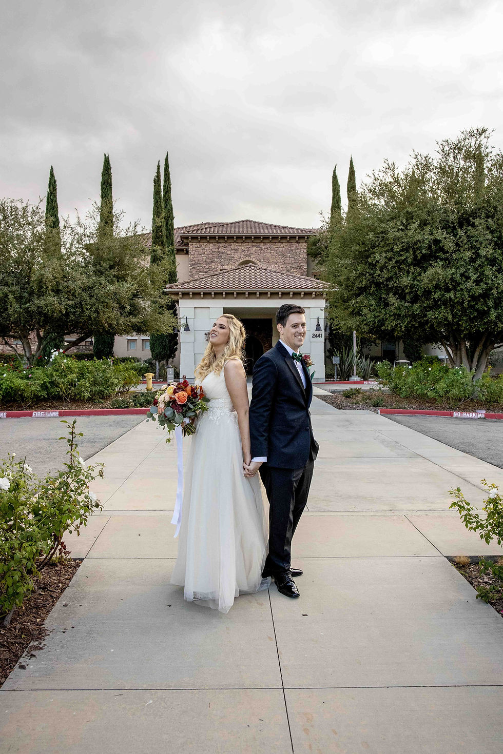 Bride and groom holding hands back-to-back, bride in a white dress with flowers. Outdoor setting with greenery and a brick building. Cloudy sky.
