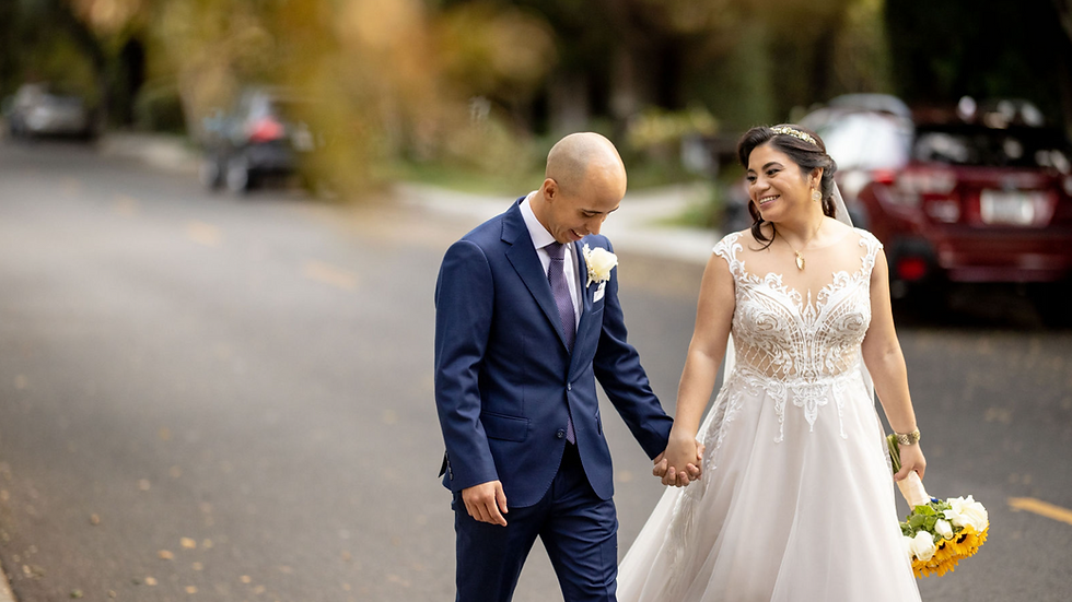 Bride in white gown and groom in blue suit happily walking down a street, holding hands. She holds a bouquet of sunflowers.