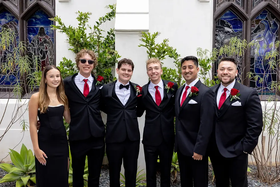 Six people in formal attire, five men in black suits with red ties and a woman in a black dress, pose smiling outside near stained glass.