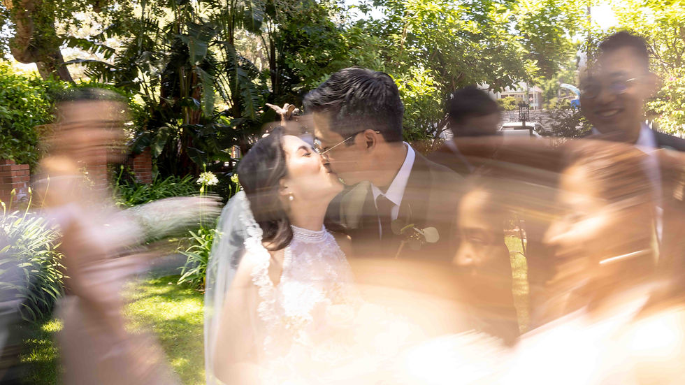 Bride and groom kiss in a garden surrounded by blurred guests, creating a dynamic, joyful atmosphere with lush greenery in the background.