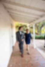 Man in a gray suit and green bow tie walks arm-in-arm with a woman in a blue dress under a white veranda. Calm, sunny setting.