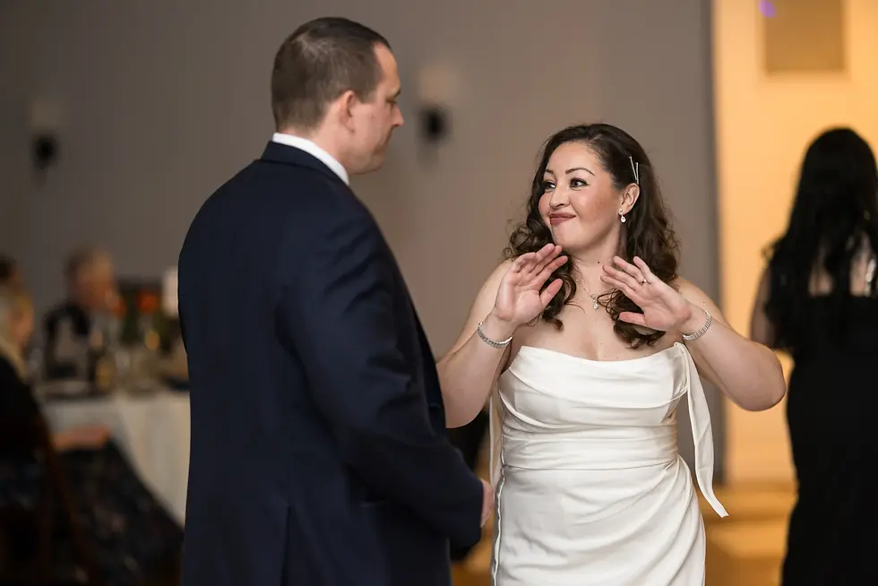 A woman in a white dress and a man in a suit dance with smiles at an indoor event, with blurred guests seated in the background. direct flash