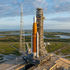 A large rocket on a launchpad at a coastal site, set against a backdrop of green fields and a blue ocean under a partly cloudy sky.