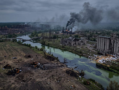 Aerial view of a bombed industrial site with black smoke, ruined buildings, scorched fields, and a river under gray skies.