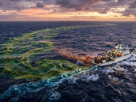 A ship sails in open ocean at sunset, creating swirling yellow patterns in the water. The sky is a mix of orange, pink, and purple hues.