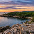 Sunset over rocky forested coast with a lighthouse, calm water, and wind turbines on distant hills.