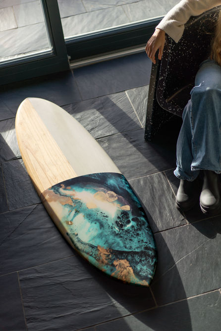 woman wearing jeans and a cream cardigan sitting in a black chair next to a wooden surfboard with artwork on a slate floor