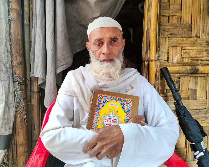 Shomsul, 68, spends much of his time reading the Quran at the entrance of his shelter. Neighbours say he rarely sleeps and prays through the night. For many Rohingya refugees, religious faith becomes a source of emotional resilience. 2021 © Sahat Zia Hero