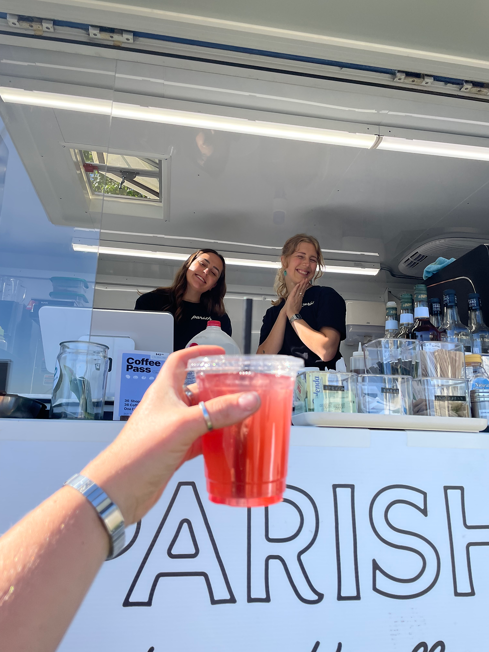 Two smiling baristas in the Parish Coffee truck who just handed a customer a yummy iced tea drink. The sun is shining and the customer is excited to try the iced drink.