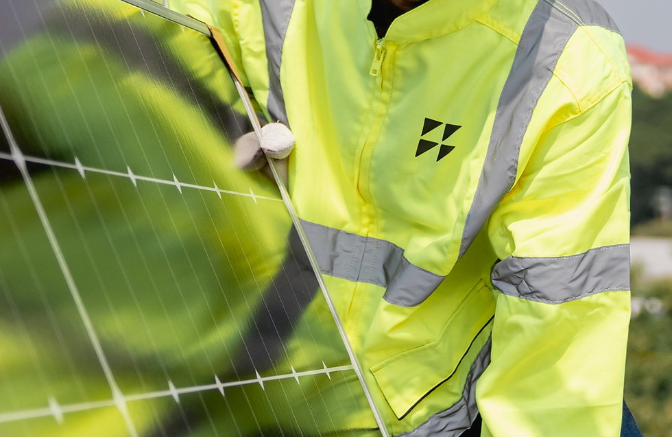 Worker in a bright yellow safety jacket installs a solar panel outdoors. The jacket has gray stripes and a black logo. Sunny day.