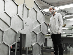 A person in a lab coat and hairnet examines a large hexagonal metal structure in an industrial setting. Text on panel reads "410 E. Cota St."