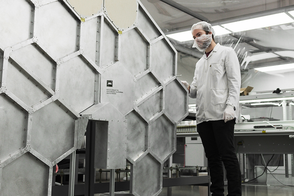 A person in a lab coat and hairnet examines a large hexagonal metal structure in an industrial setting. Text on panel reads "410 E. Cota St."