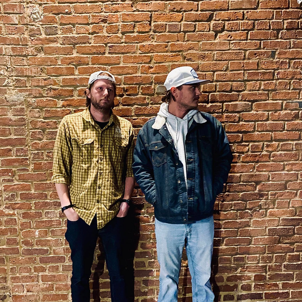 Two men in casual wear standing against a brick wall.