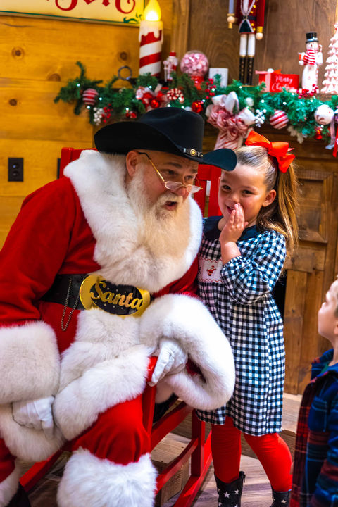 Girl whispering to Santa wearing a cowboy hat.