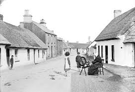old lady sat at spinning wheel outside a row of cottages and 2 children next to her, old black and white photo