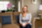 Stacy sitting on a beige sofa holding a white mug and smiling at the camera