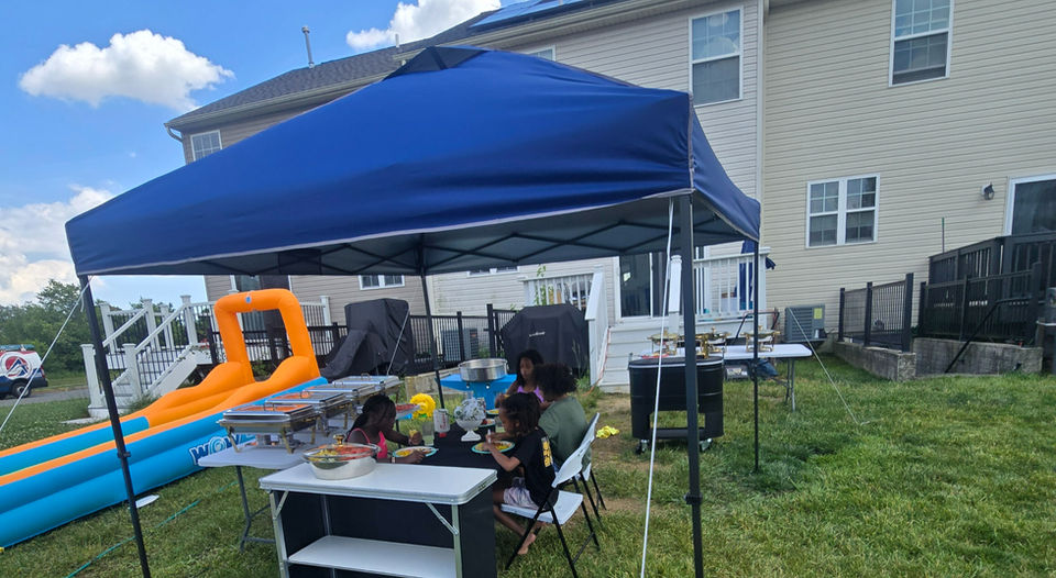 Children eat under a blue canopy tent in a backyard.