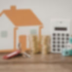 House model with stacks of coins, keys, and calculator on a wooden table, symbolizing home finance. Blocks show numbers, creating a planning mood.
