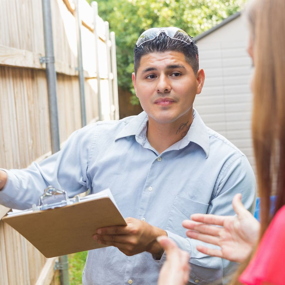 Man with clipboard talks to a person outdoors near a wooden fence, wearing a light blue shirt and safety glasses, in a professional manner.