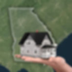 A hand holds a small gray house over a map of Georgia. The image evokes a sense of home ownership. Green landscape background.