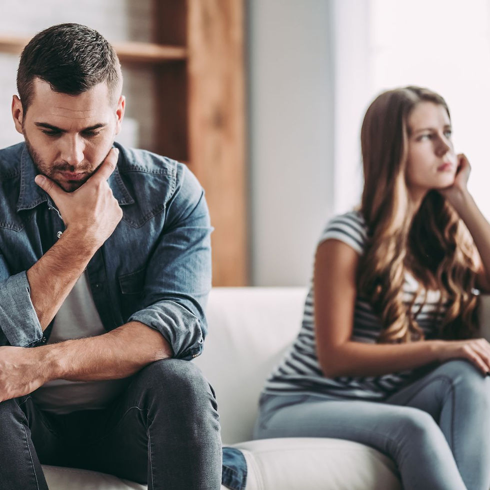 A man and a woman sit apart on a sofa, looking away from each other with thoughtful expressions. The setting is a cozy living room.