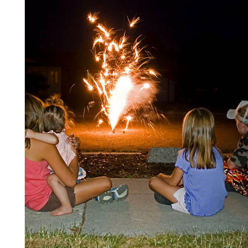 Children sitting on a sidewalk at night, watching bright fireworks. One child is held by an adult. Warm and festive atmosphere.