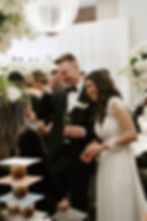 Bride and groom laughing with a guest at a wedding reception. They're dressed formally. Elegant decor with white flowers in the background.