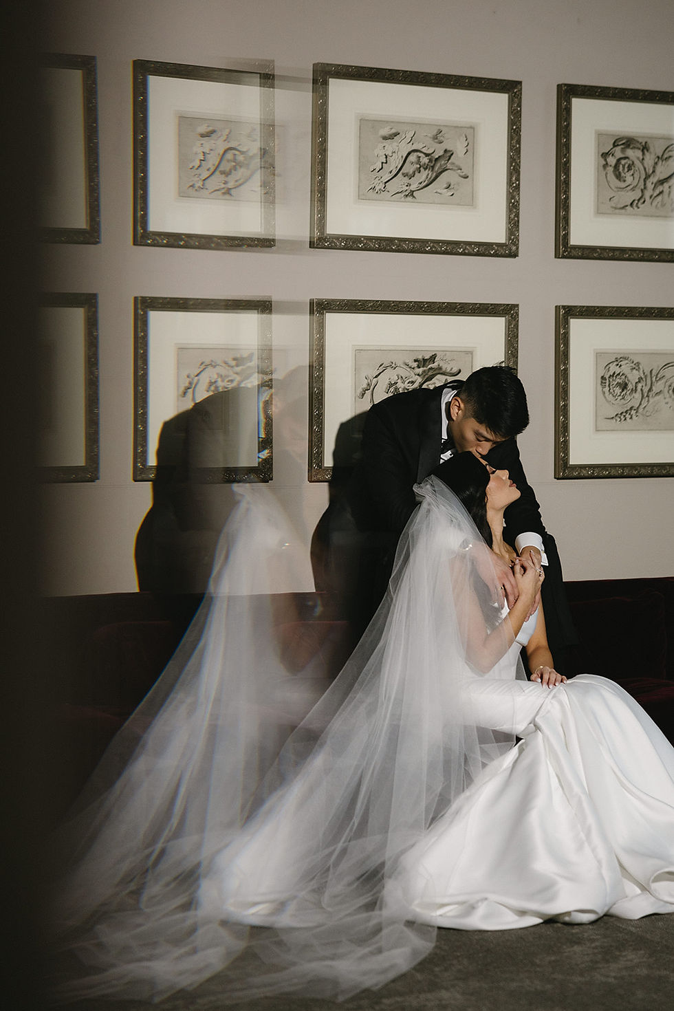 Bride in white gown and veil sits as groom in black tuxedo leans to kiss her forehead. Art frames on wall. Romantic and intimate scene.