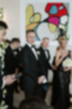 Groom in black suit with groomsmen, a woman holding flowers, colorful abstract art in background. Formal, emotional wedding scene.