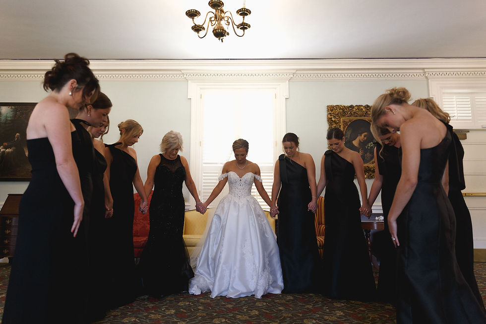 Bride in white gown and bridesmaids in black dresses hold hands in a circle, bowing heads in a decorated room with chandeliers.